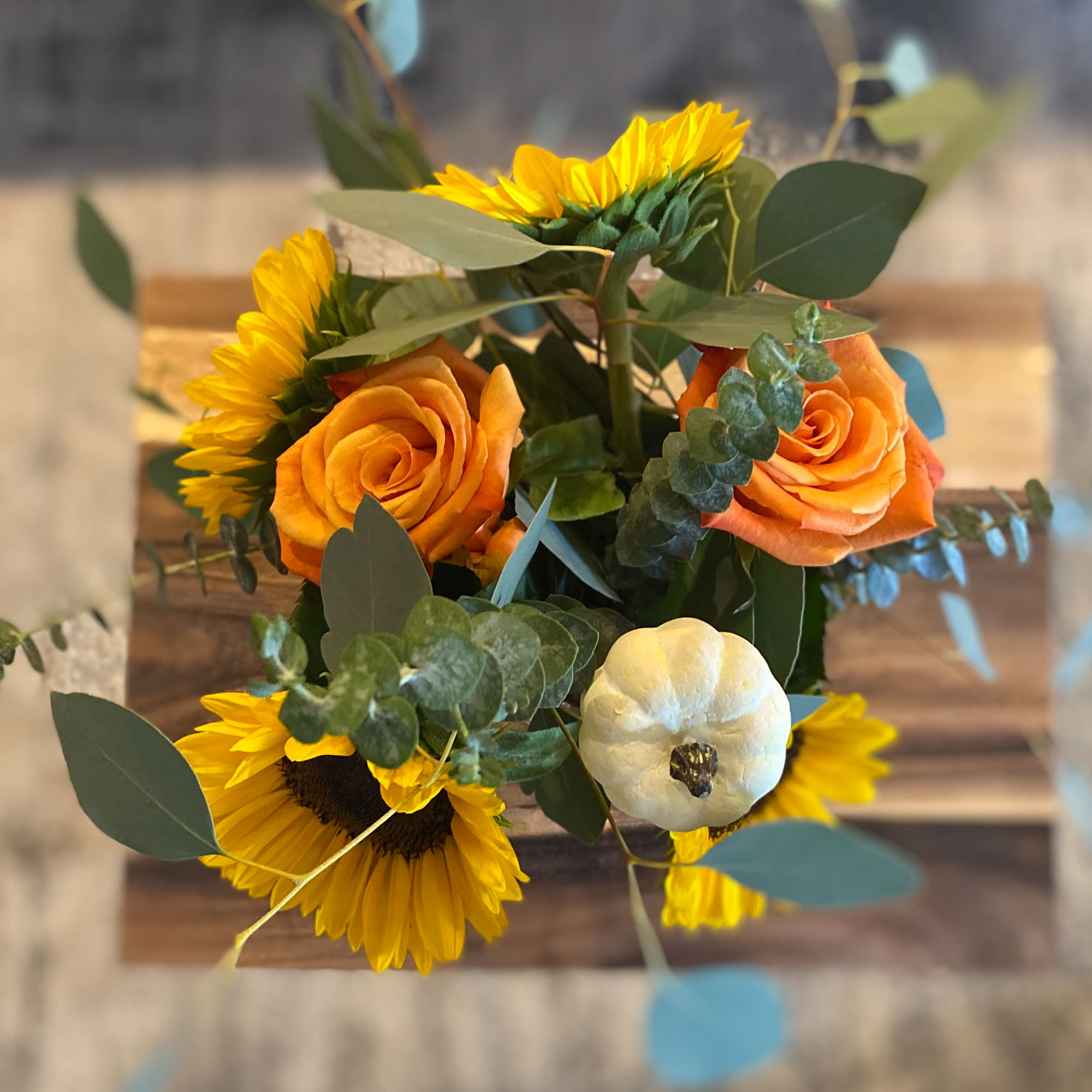Overhead shot of a flower arrangement in a glass vase on a wooden coffee table, with sunflowers and pumpkins.