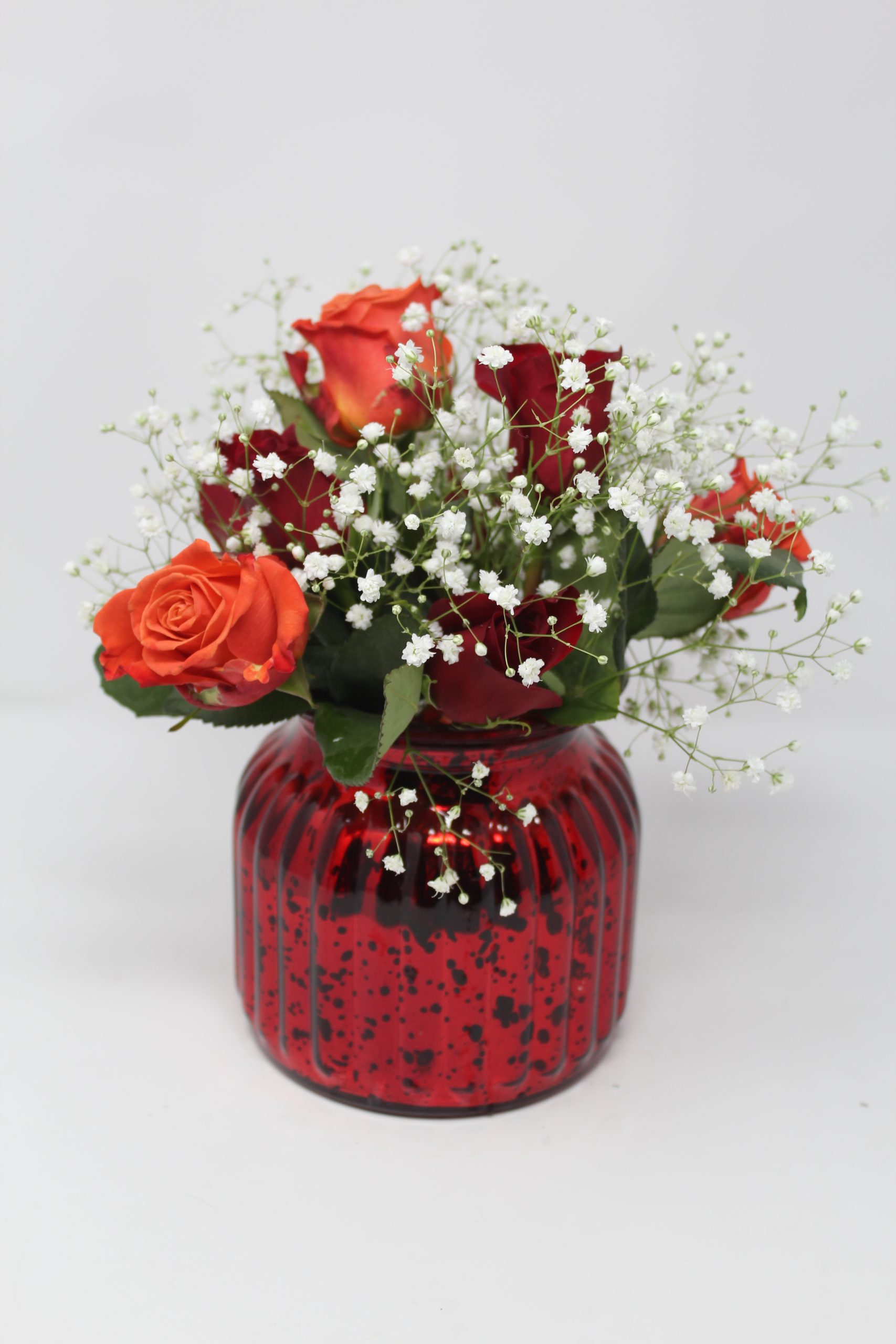 Photo of a small red vase with orange and red roses, and other flowers.